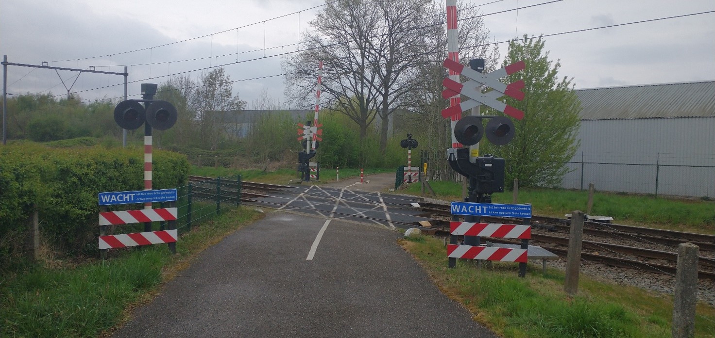 Bewaakte spoorwegovergang met aan de zijkanten gras, enkele bomen en in de verte gebouwen van het bedrijventerrein 